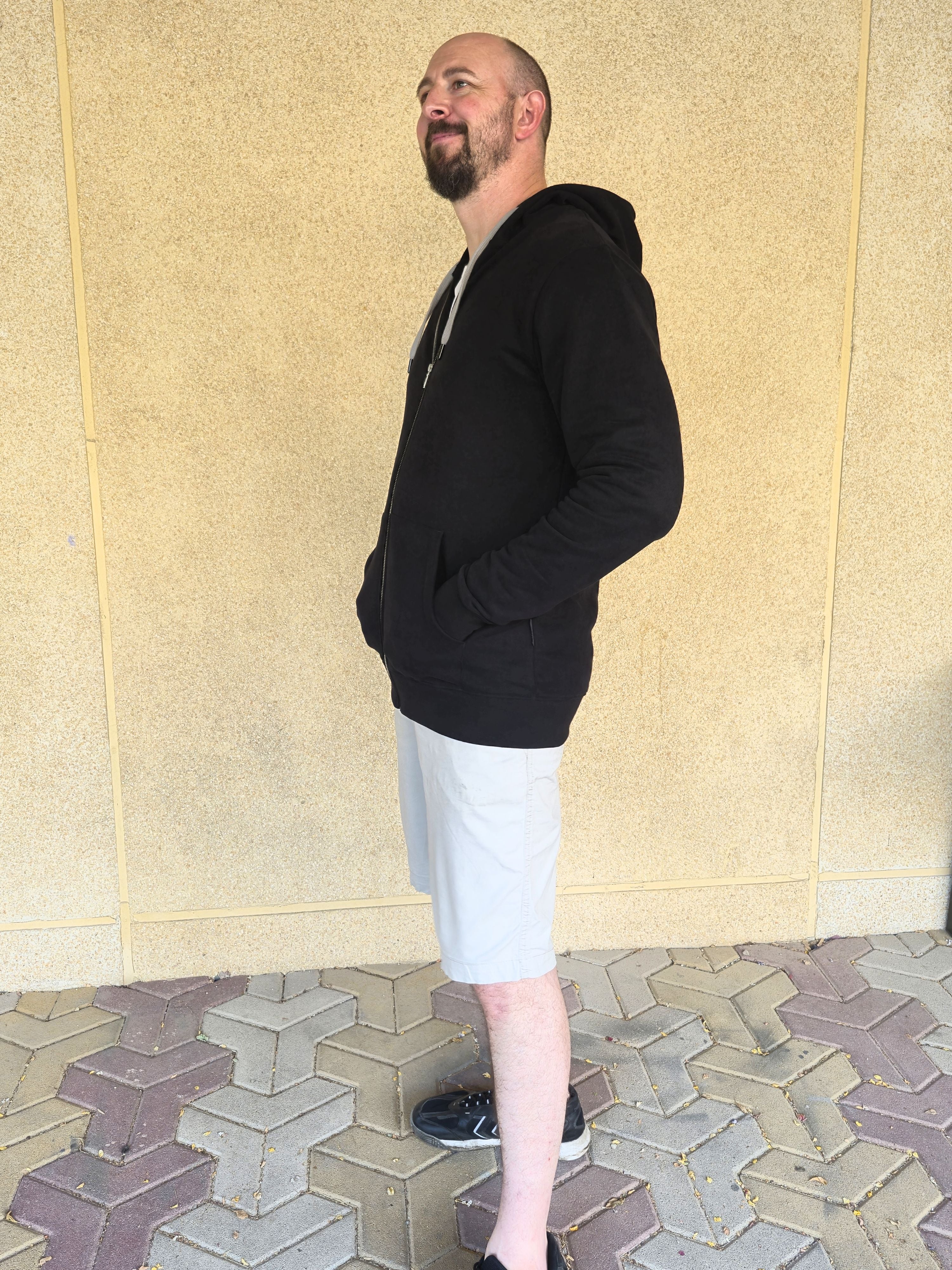 A side-on view of a tall and athletic guy wearing an XL tall black hoodie, standing in front of a beige wall, making hands in pockets and smiling.