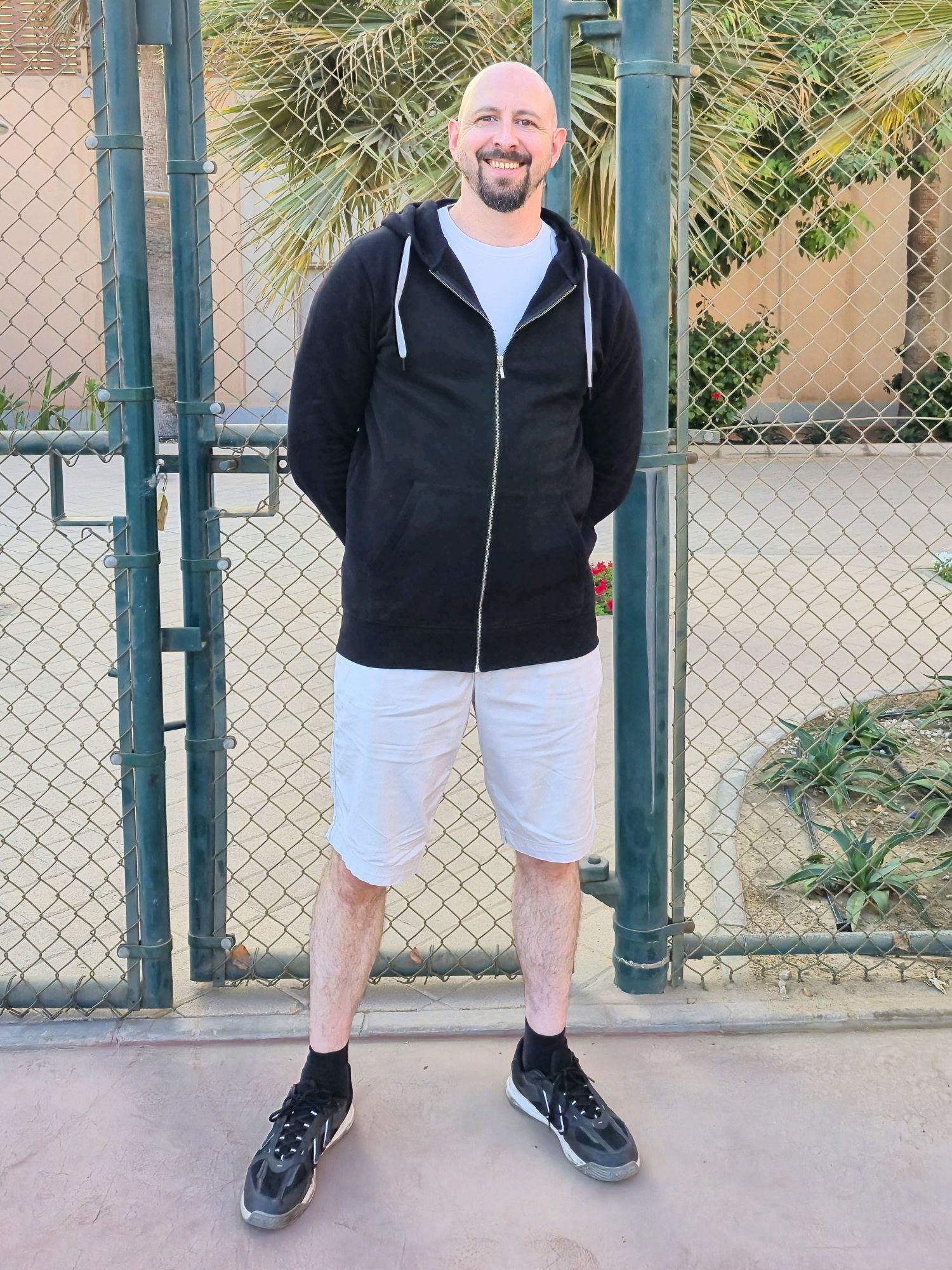 A tall and athletic guy wearing an XL tall black hoodie, standing in a tennis court, hands behind back and smiling.