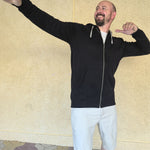 A tall slim man wearing a tall black full zip hoodie and striking a super star pose and laughing.
