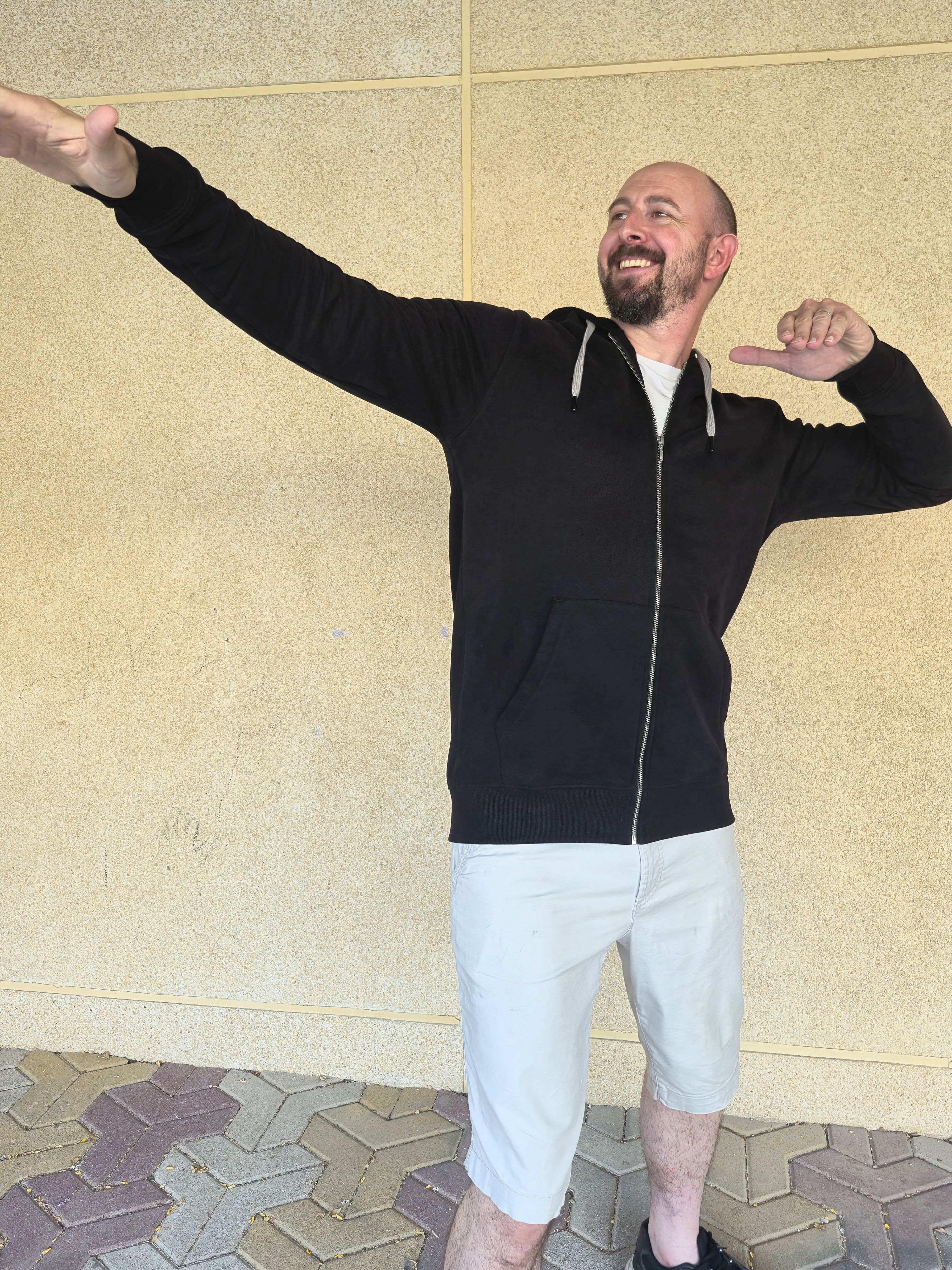 A tall slim man wearing a tall black full zip hoodie and striking a super star pose and laughing.