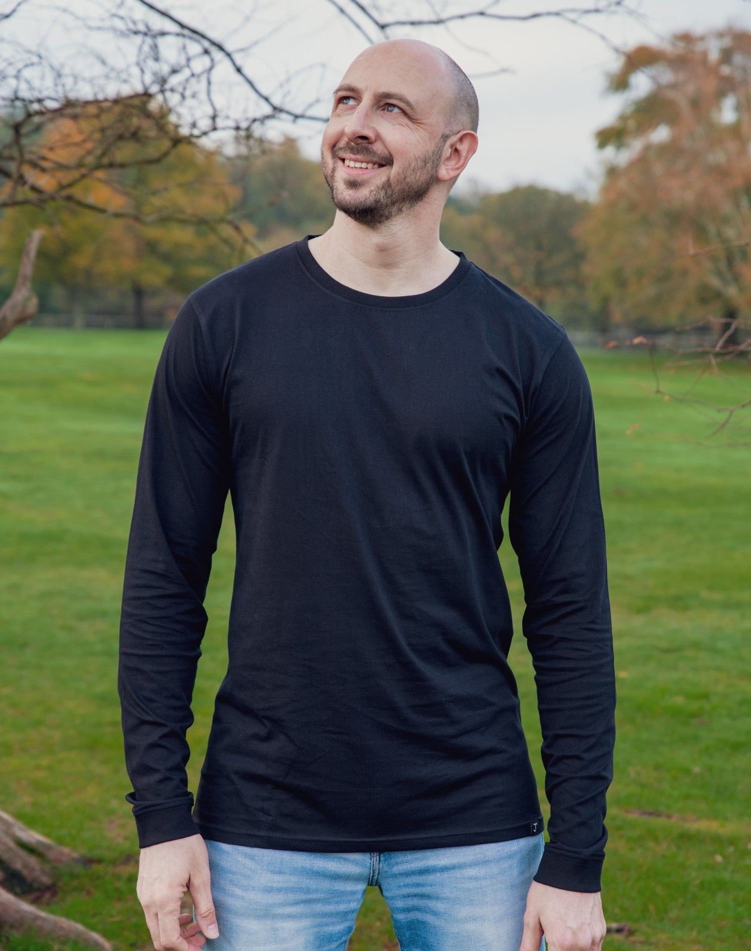 A tall and slim man standing in a park. The smiling model is wearing a black long-sleeve tall t-shirt. The tall black long-sleeve t-shirt features a 2.5" longer sleeves, 100% organic cotton, and is soft & preshrunk. The black long-sleeve t-shirt is ideal for tall slim men 6'2"+.