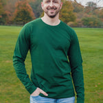 A tall and slim man standing in a park with one hand in his pocket. The smiling model is wearing a dark green long-sleeve tall t-shirt. The tall dark green long-sleeve t-shirt features a 2.5" longer sleeves, 100% organic cotton, and is soft & preshrunk. The dark green long-sleeve t-shirt is ideal for tall slim men 6'2"+.