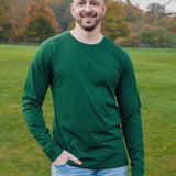 A tall and slim man standing in a park with one hand in his pocket. The smiling model is wearing a dark green long-sleeve tall t-shirt. The tall dark green long-sleeve t-shirt features a 2.5" longer sleeves, 100% organic cotton, and is soft & preshrunk. The dark green long-sleeve t-shirt is ideal for tall slim men 6'2"+.