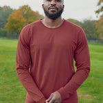 A tall and slim man standing in a park. The smiling model is wearing a dark orange long-sleeve tall t-shirt. The tall dark orange long-sleeve t-shirt features a 2.5" longer sleeves, 100% organic cotton, and is soft & preshrunk. The dark orange long-sleeve t-shirt is ideal for tall slim men 6'2"+.