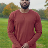 A tall and slim man standing in a park. The smiling model is wearing a dark orange long-sleeve tall t-shirt. The tall dark orange long-sleeve t-shirt features a 2.5" longer sleeves, 100% organic cotton, and is soft & preshrunk. The dark orange long-sleeve t-shirt is ideal for tall slim men 6'2"+.
