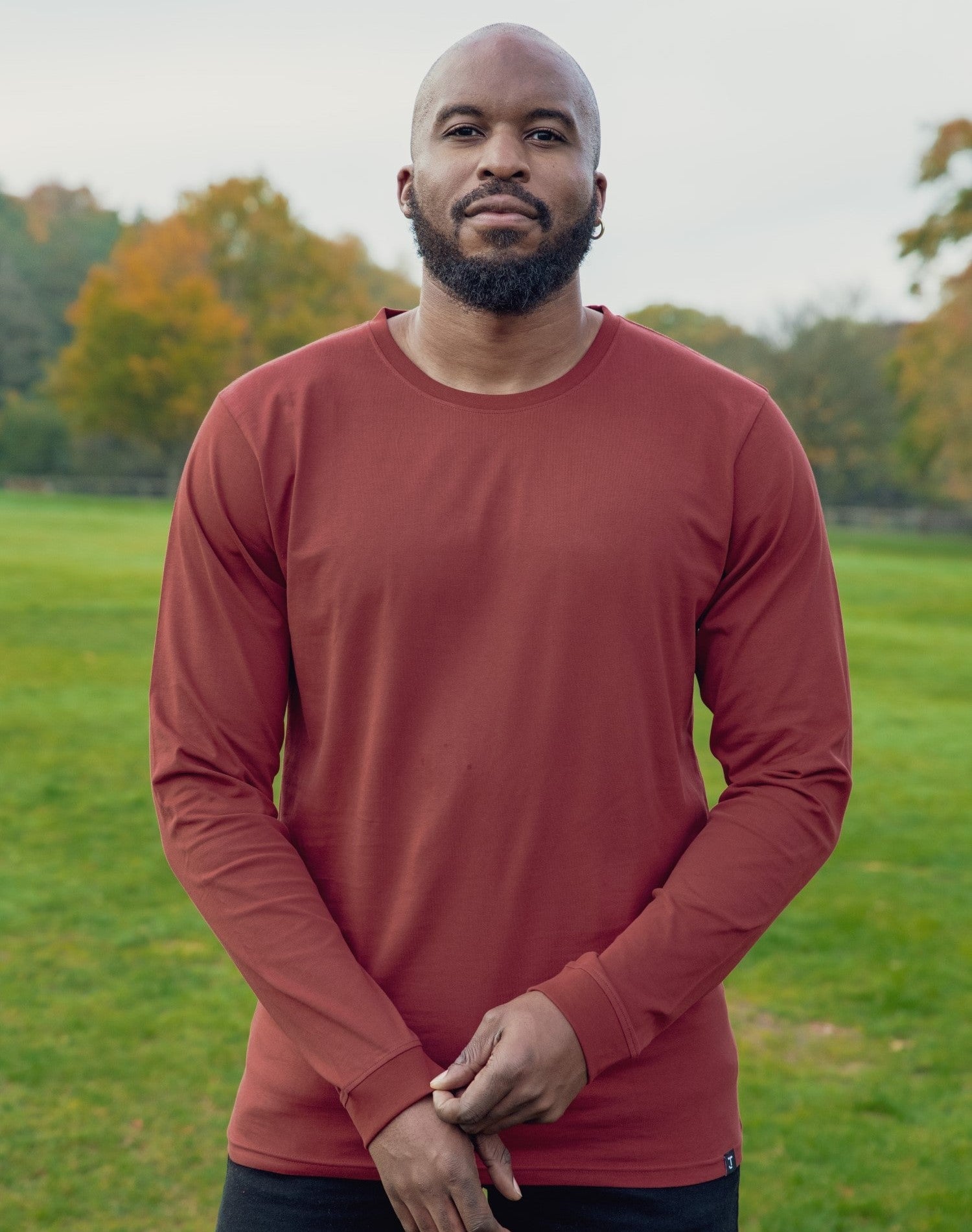 A tall and slim man standing in a park. The smiling model is wearing a dark orange long-sleeve tall t-shirt. The tall dark orange long-sleeve t-shirt features a 2.5" longer sleeves, 100% organic cotton, and is soft & preshrunk. The dark orange long-sleeve t-shirt is ideal for tall slim men 6'2"+.