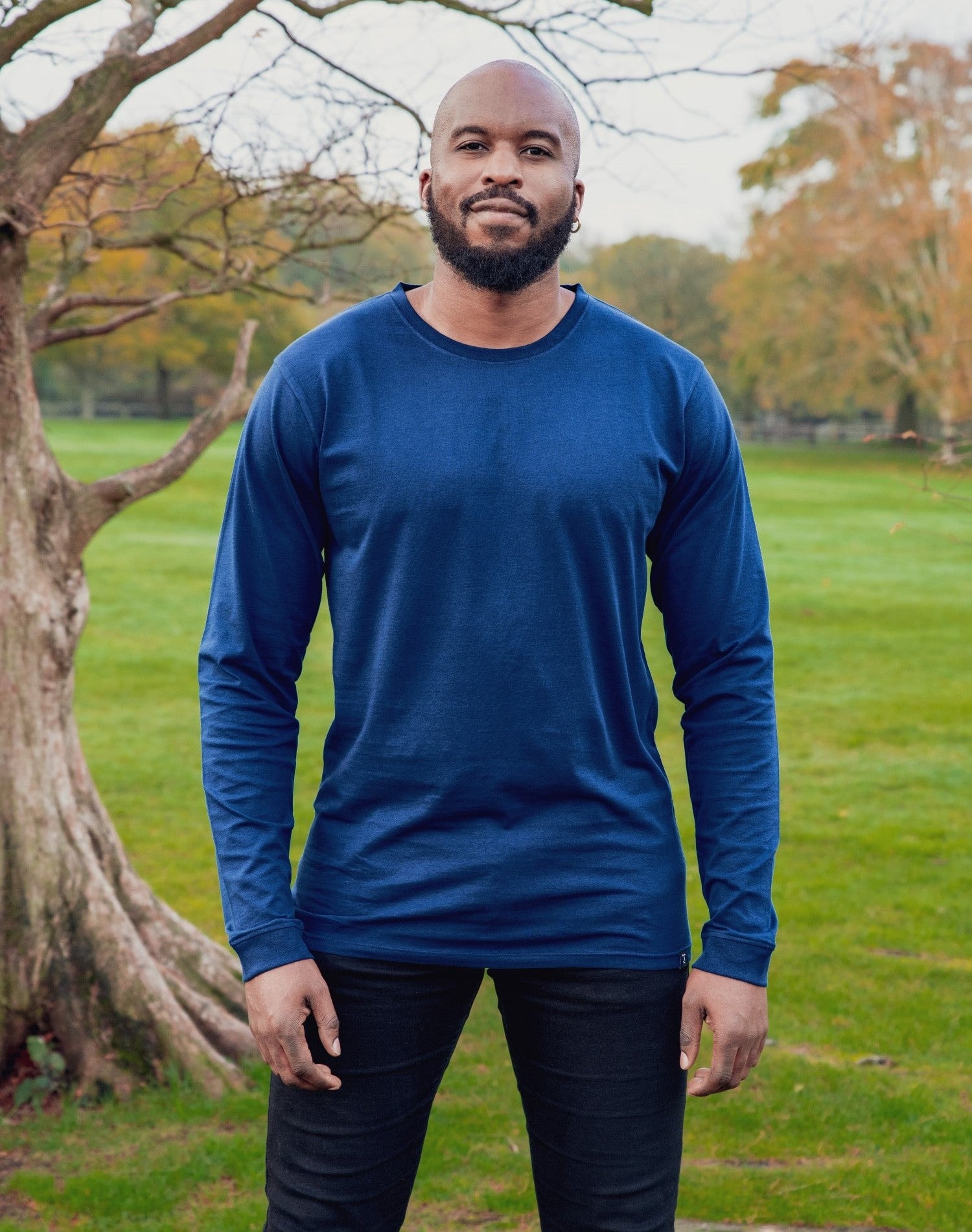 A tall and slim man standing in a park. The smiling model is wearing a navy blue long-sleeve tall t-shirt. The tall navy blue long-sleeve t-shirt features a 2.5" longer sleeves, 100% organic cotton, and is soft & preshrunk. The navy blue long-sleeve t-shirt is ideal for tall slim men 6'2"+.