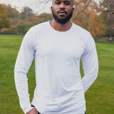 A tall and slim man standing in a park with one hand in his pocket. The smiling model is wearing a white long-sleeve tall t-shirt. The tall white long-sleeve t-shirt features a 2.5" longer sleeves, 100% organic cotton, and is soft & preshrunk. The white long-sleeve t-shirt is ideal for tall slim men 6'2"+.