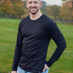 A tall athletic guy wearing a long sleeve black tall t-shirt and smiling in a park with hand in back pocket.