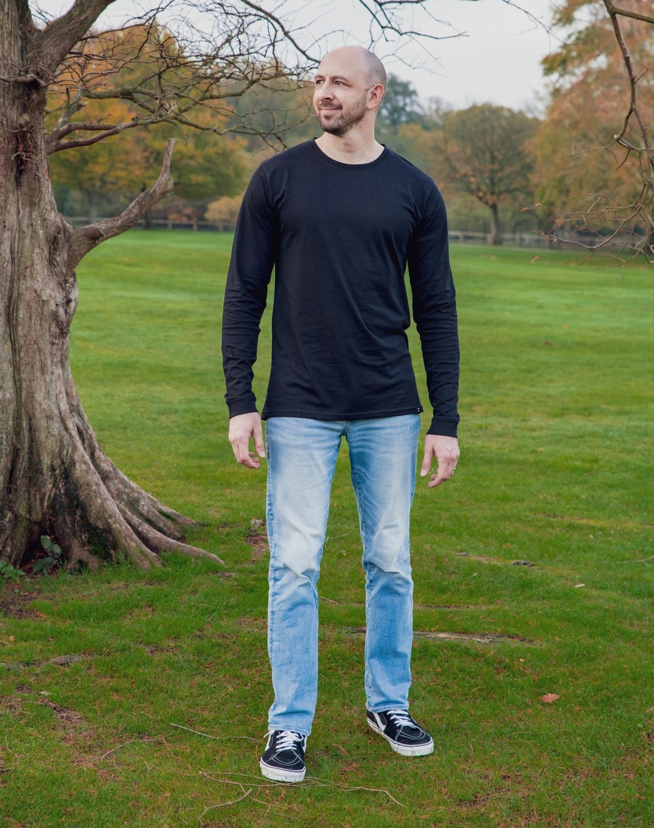 A head to toe shot of a tall athletic guy in a park wearing a black long sleeve tall t-shirt, looking to the right