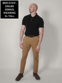 Thumbnail for A head to toe shot of a tall slim guy wearing a black XL tall polo shirt tucked into a brown belt.