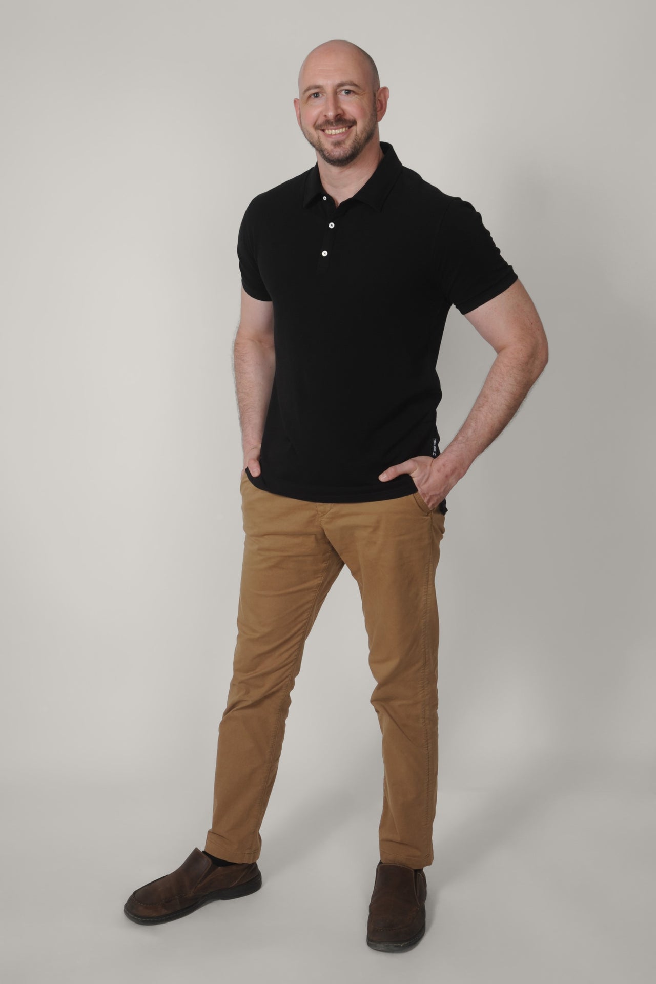 A head to toe shot of a tall skinny guy wearing an XL tall black pique polo shirt, hands in pockets.