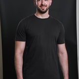 A tall and slim guy smiling in the studio and wearing a charcoal XL tall slim t-shirt.