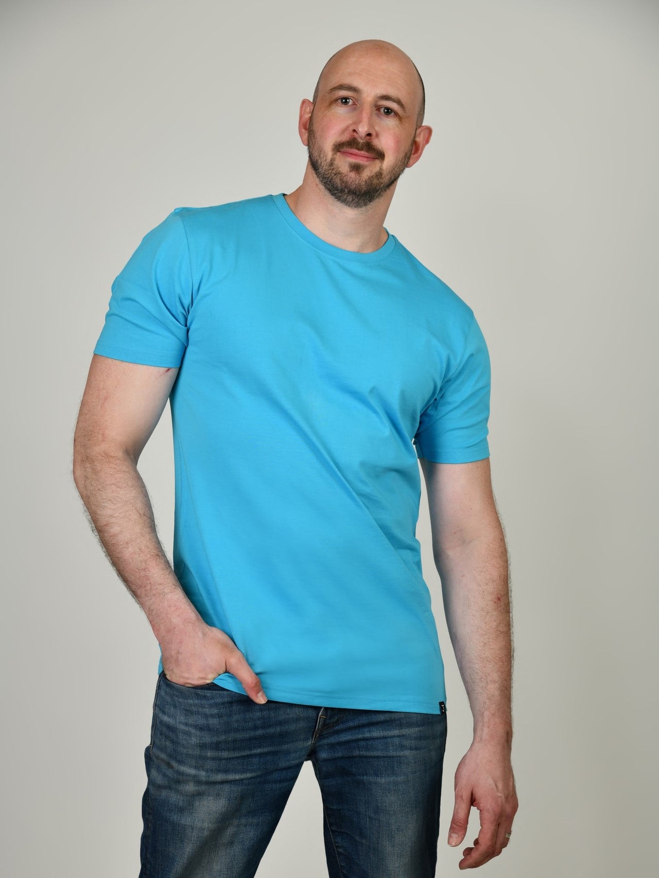 A tall and slim guy in the studio, one hand in pocket and wearing a cyan XL tall slim t-shirt.