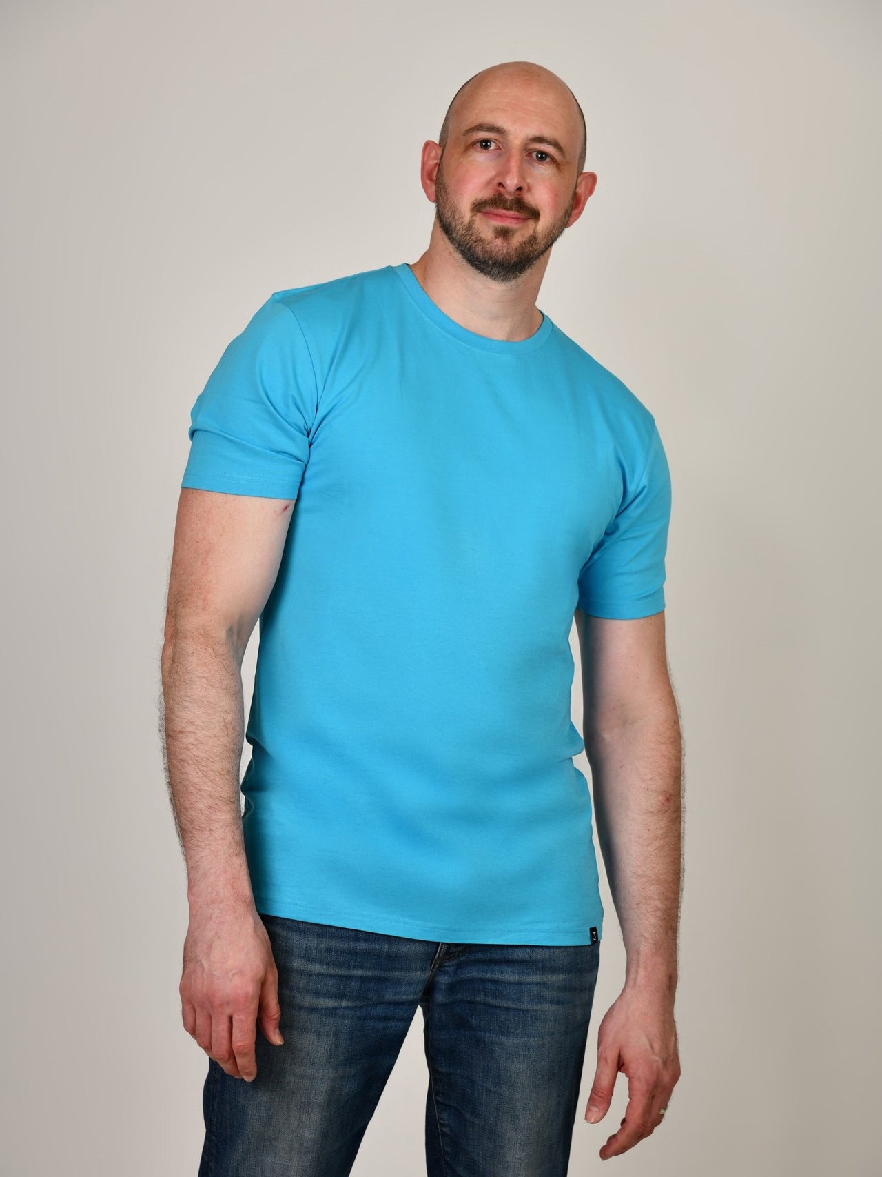 A tall and slim guy smiling in the studio and wearing a cyan XL tall slim t-shirt.