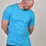 A tall and slim guy in the studio, hands behind back and wearing a cyan XL tall slim fit t-shirt.