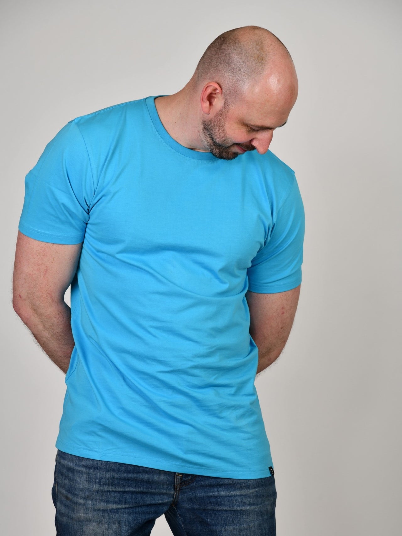 A tall and slim guy in the studio, hands behind back and wearing a cyan XL tall slim fit t-shirt.
