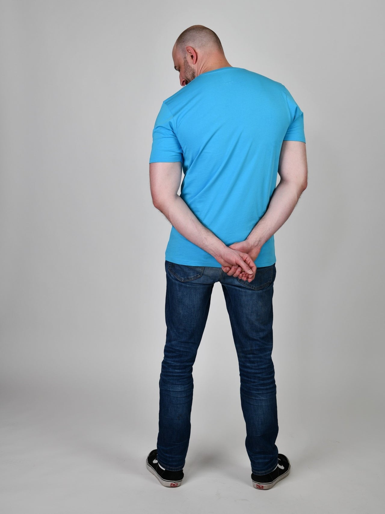 A shot from behind of a tall and slim guy in the studio and wearing a cyan XL tall slim t-shirt.
