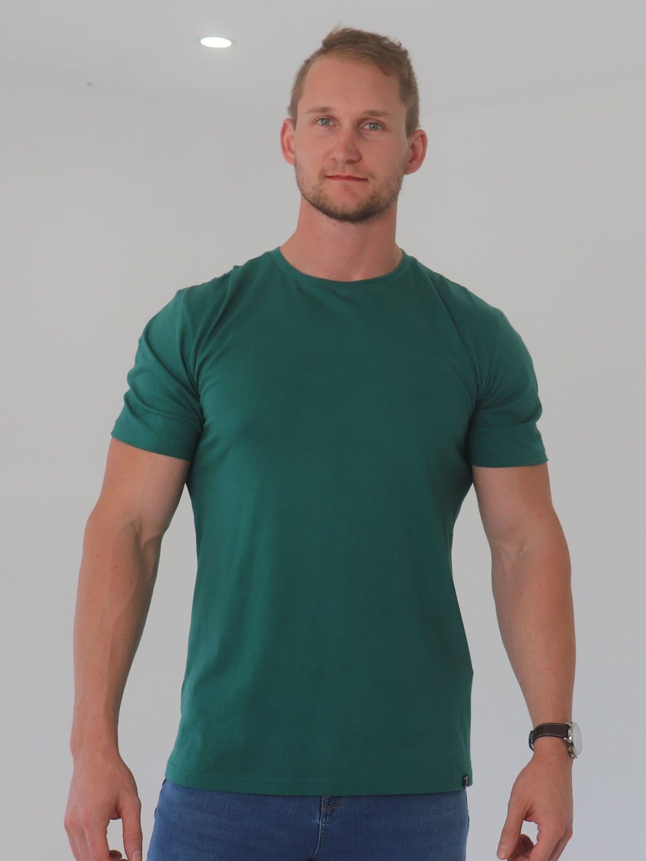 A tall and broad guy smiling in the studio and wearing a dark green 2XL tall slim t-shirt.