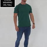 A head to toe shot of a tall and broad guy in the studio wearing a dark green 2XL tall slim t-shirt.