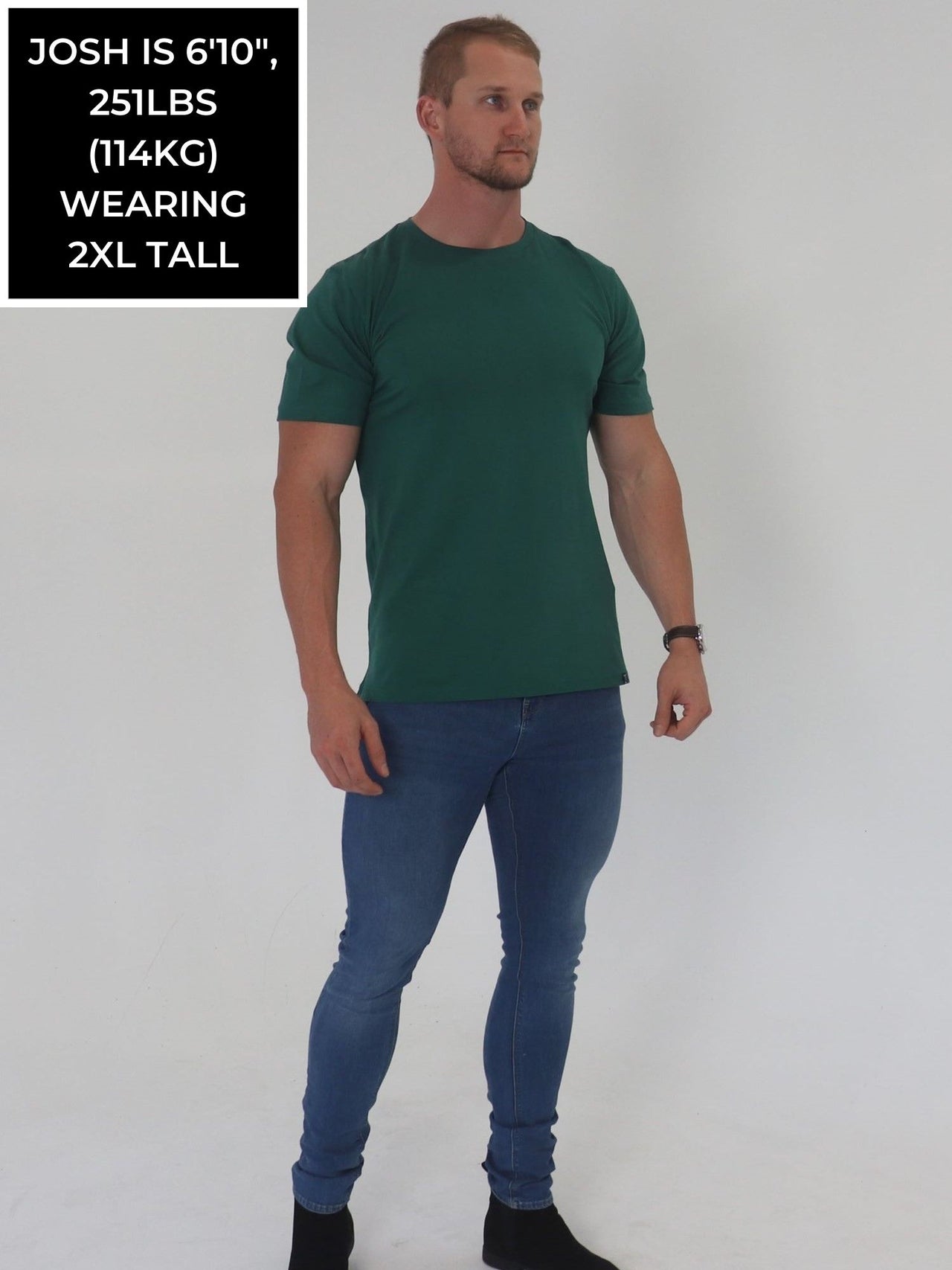 A head to toe shot of a tall and broad guy in the studio wearing a dark green 2XL tall slim t-shirt.