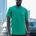 A tall and skinny guy in the street and wearing a green minimal graphic tall t-shirt.