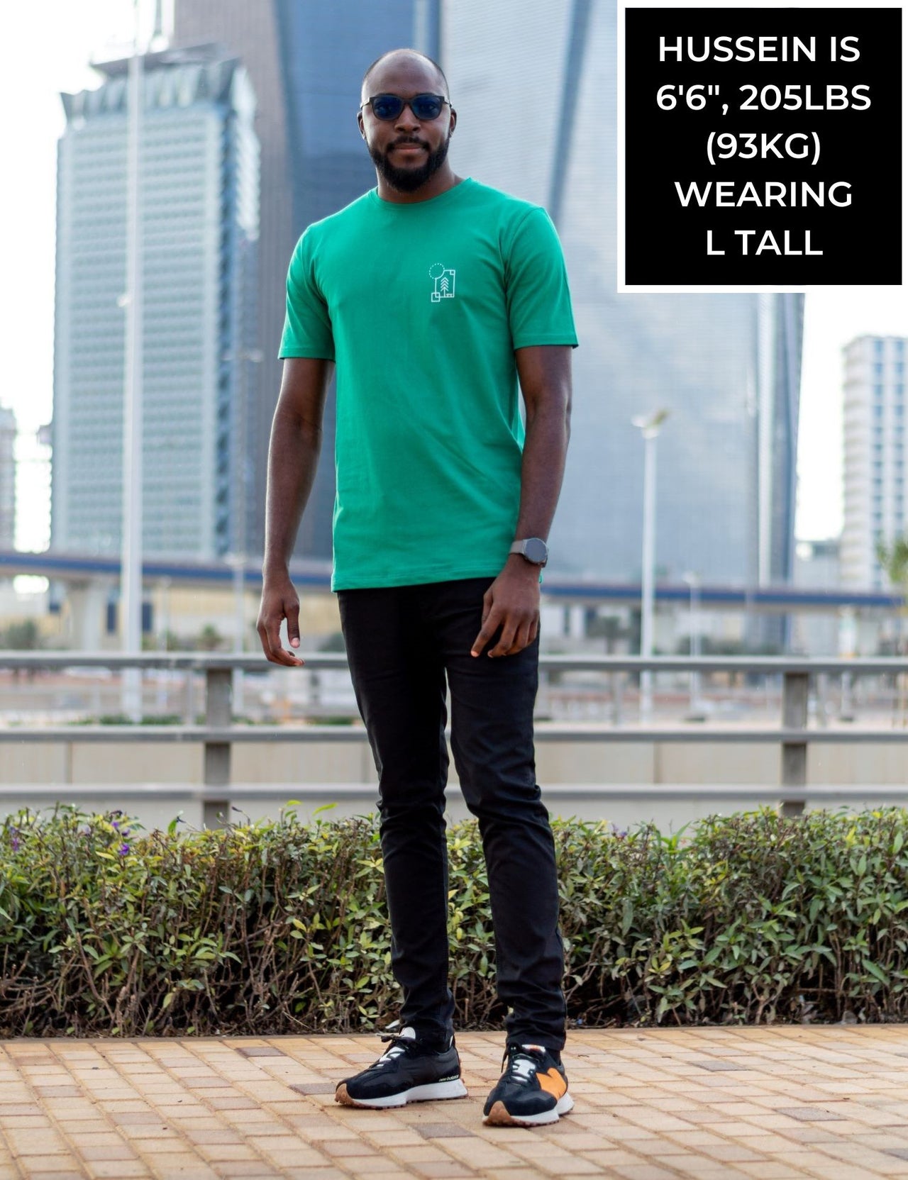 A head to toe shot of a tall and skinny guy in the street and wearing a green minimal graphic tall t-shirt.