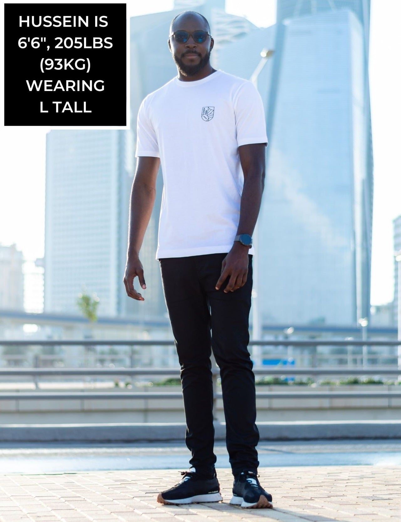 A head to toe shot of a tall and skinny guy in the street and wearing a white minimal graphic tall t-shirt.