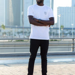 A head to toe shot of a tall and skinny guy in the street, arms folded, and wearing a white minimal graphic tall t-shirt.