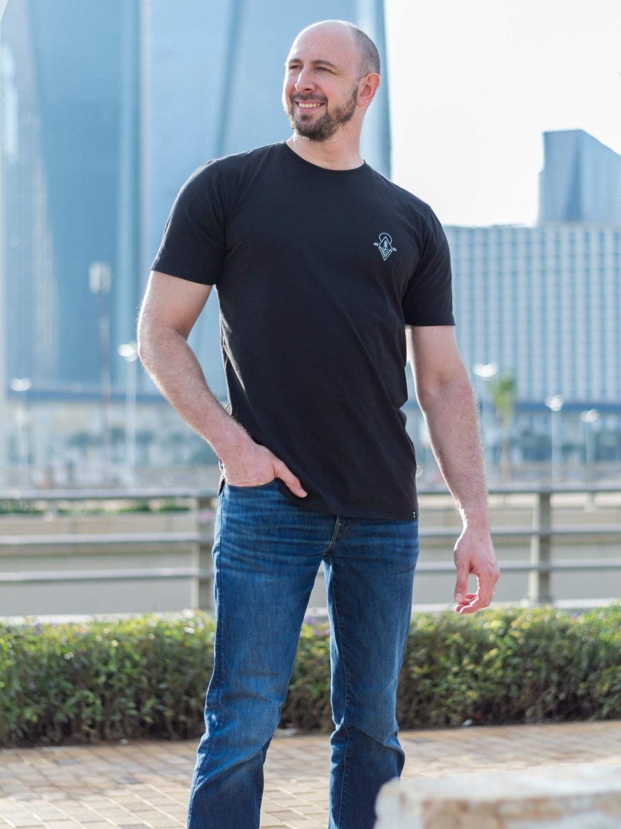 A tall and slim guy in the street, one hand in his pocket and wearing a black minimal graphic tall t-shirt.