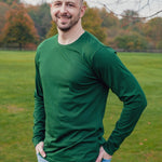 A tall athletic guy wearing a long sleeve dark green tall t-shirt and smiling in a park with hands in both pockets.