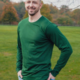 A tall athletic guy wearing a long sleeve dark green tall t-shirt and smiling in a park with hands in both pockets.