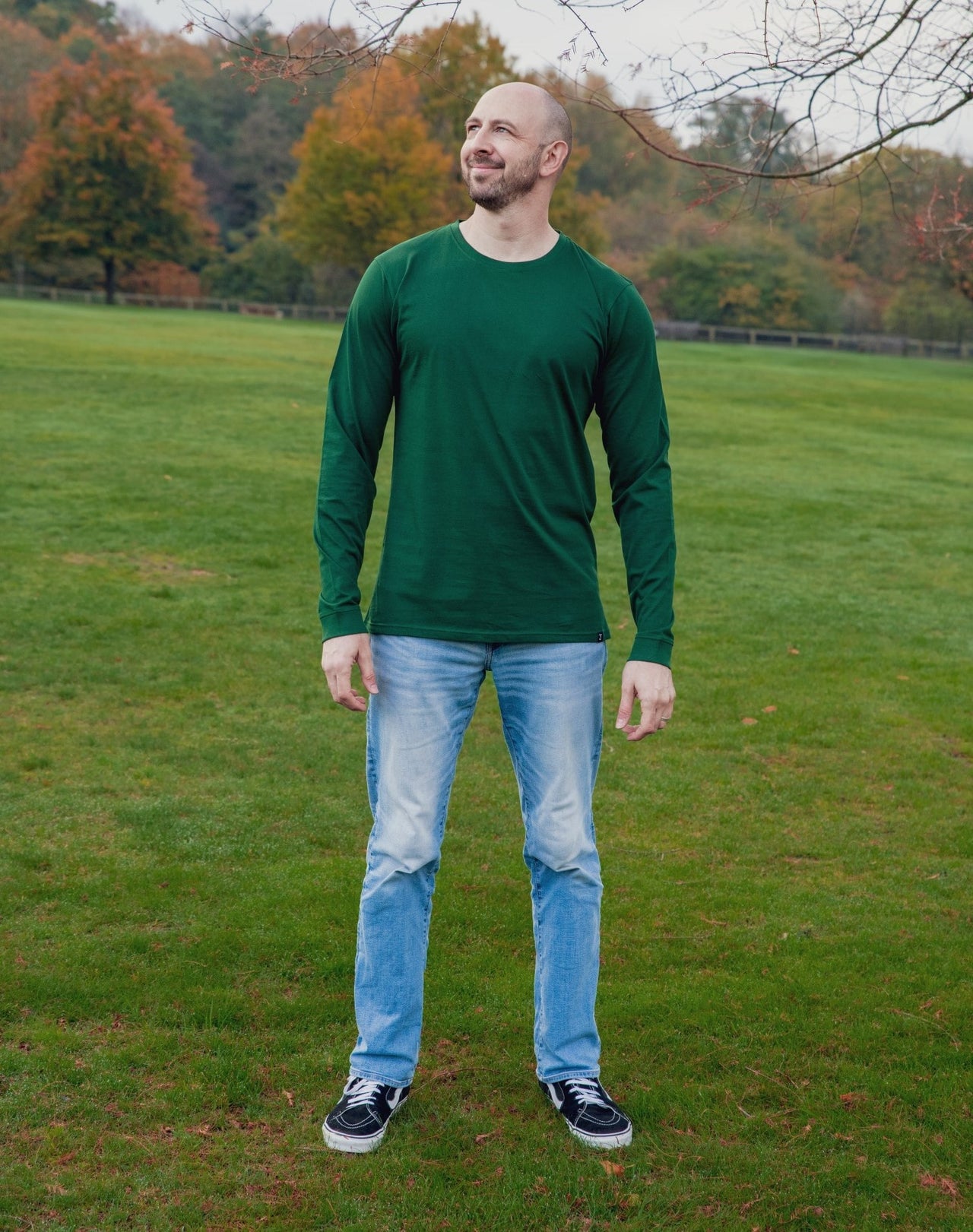 A head to toe shot of a tall athletic guy in a park, looking up at the sky and wearing a dark green long sleeve tall t-shirt.