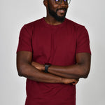 A tall and slim guy smiling in the studio, hands folded and wearing a cabernet L tall slim t-shirt.