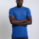 A tall and athletic guy with hands folded in the studio wearing a blue large tall slim t-shirt.