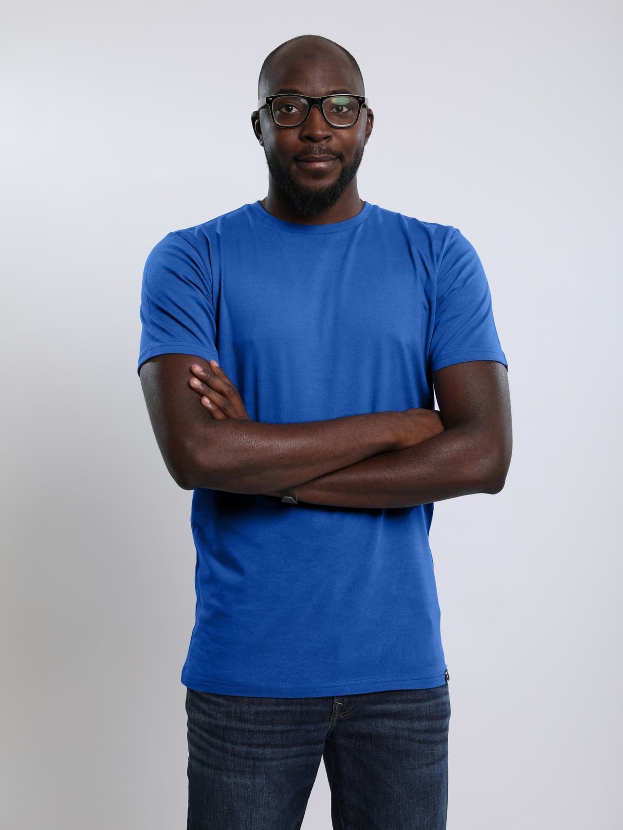 A tall and athletic guy with hands folded in the studio wearing a blue large tall slim t-shirt.