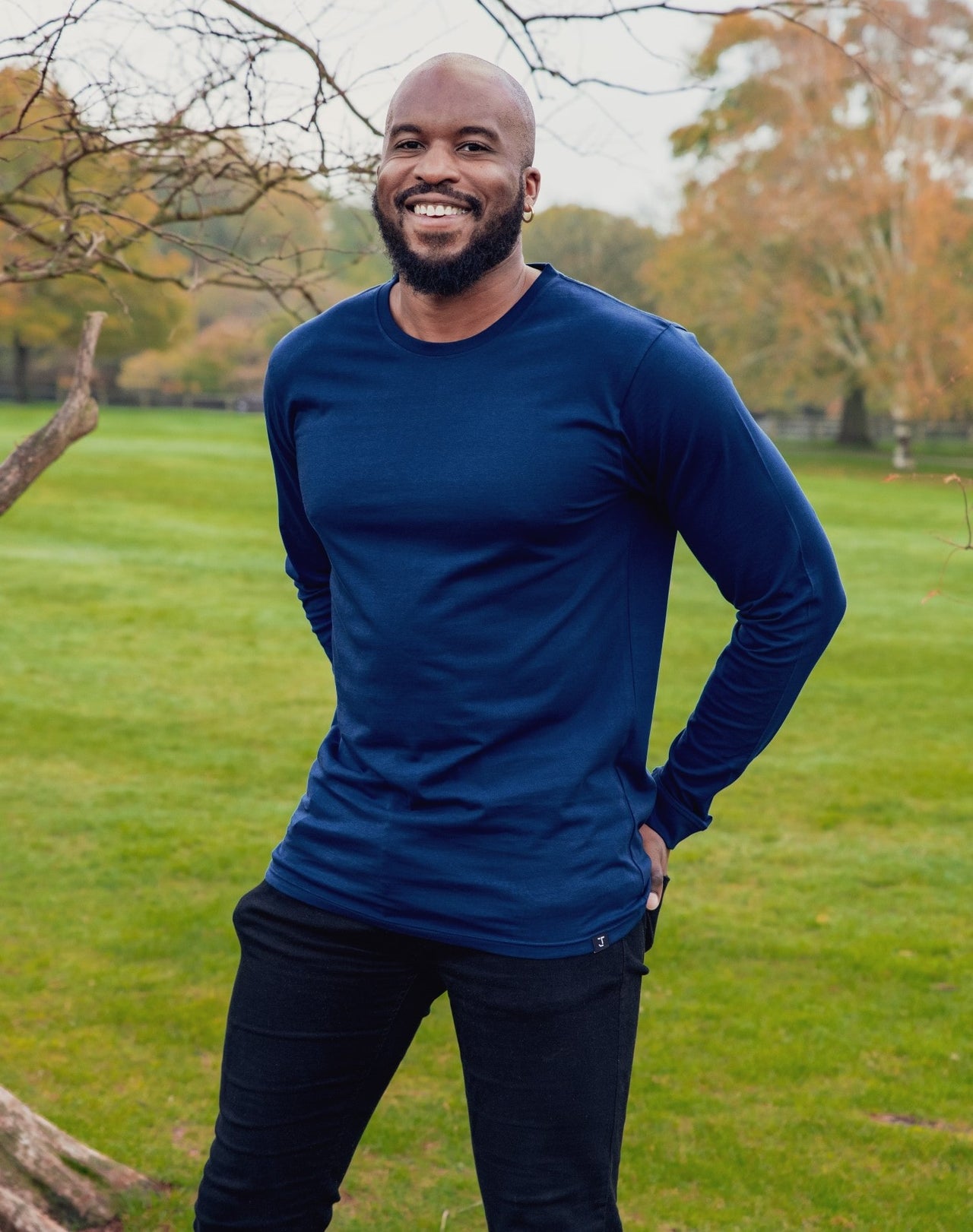 A tall athletic guy wearing a long sleeve navy tall t-shirt and smiling in a park with hands behind back.