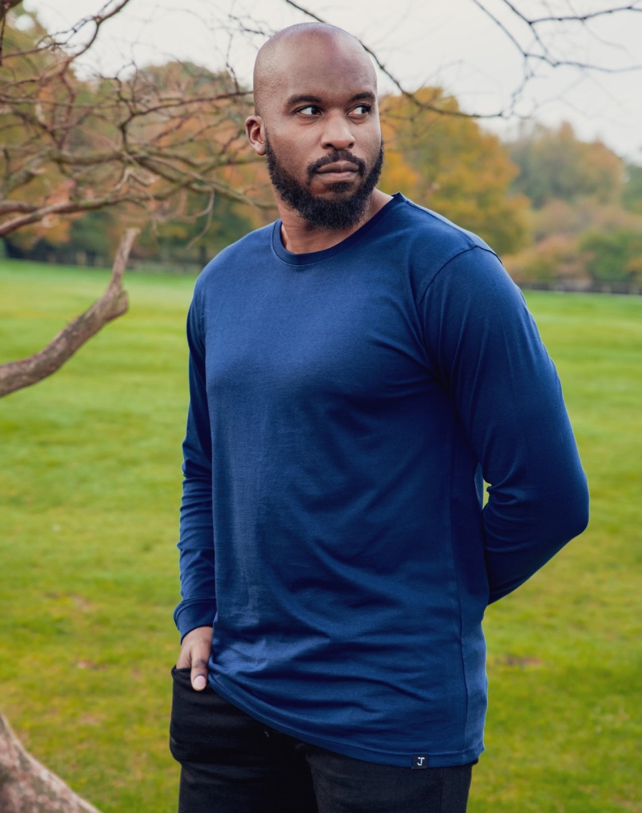 A tall athletic guy wearing a long sleeve navy tall t-shirt in a park with one hand in pocket.