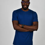 A tall and slim guy in the studio, hands folded and wearing a navy blue L tall slim t-shirt.