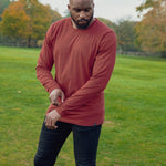 A shot of a tall athletic guy in a park wearing a dark orange long sleeve tall t-shirt, looking down at the t-shirt logo.