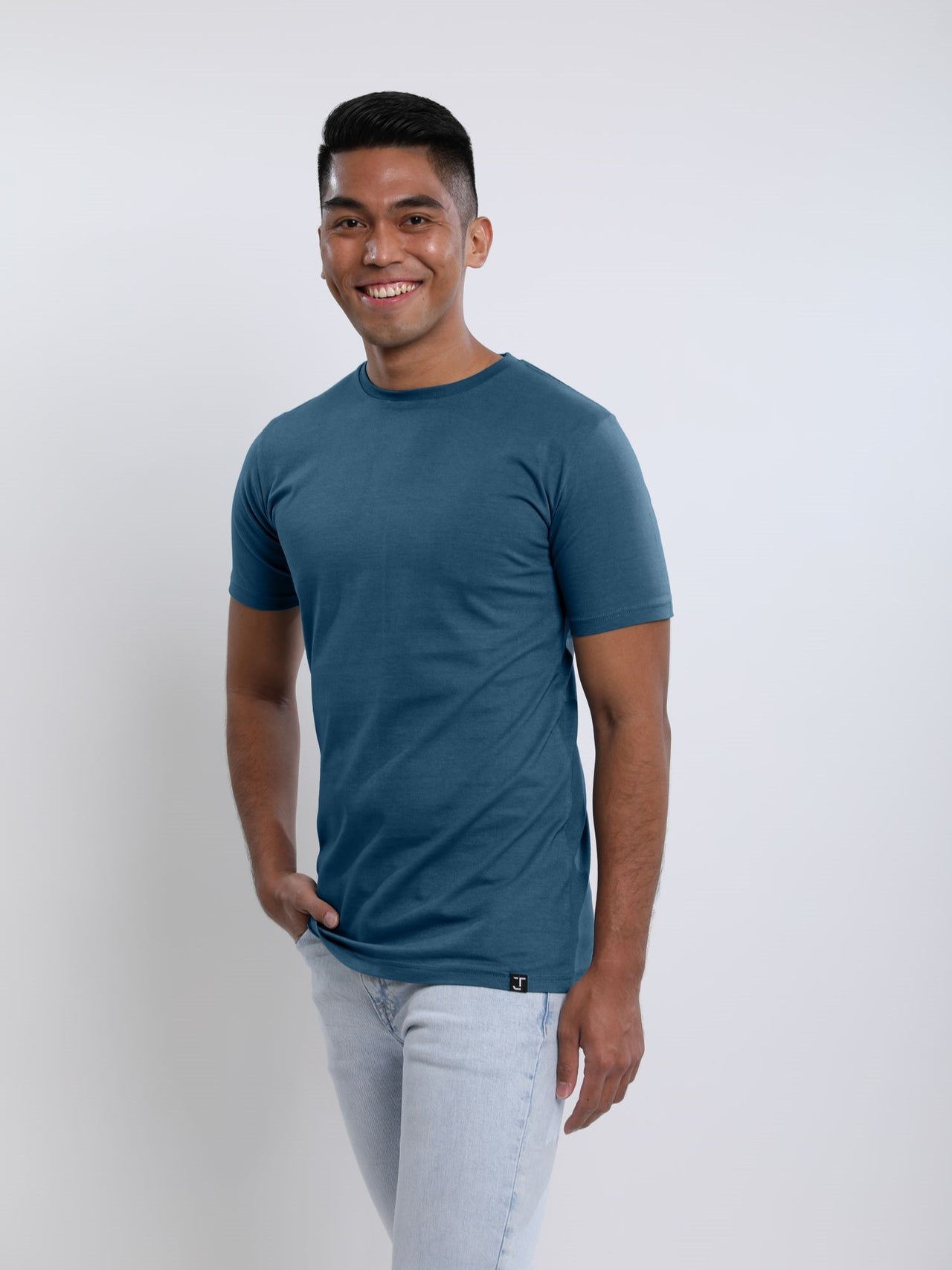 An upper body shot of a tall skinny guy wearing a petrol medium tall t-shirt, hand in pocket and smiling.