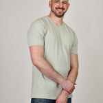 A tall and slim guy smiling in the studio, one hand grasping the other arm and wearing a sage green XL tall slim v-neck t-shirt.