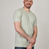 A tall and slim guy smiling in the studio, one hand grasping the other arm and wearing a sage green XL tall slim v-neck t-shirt.