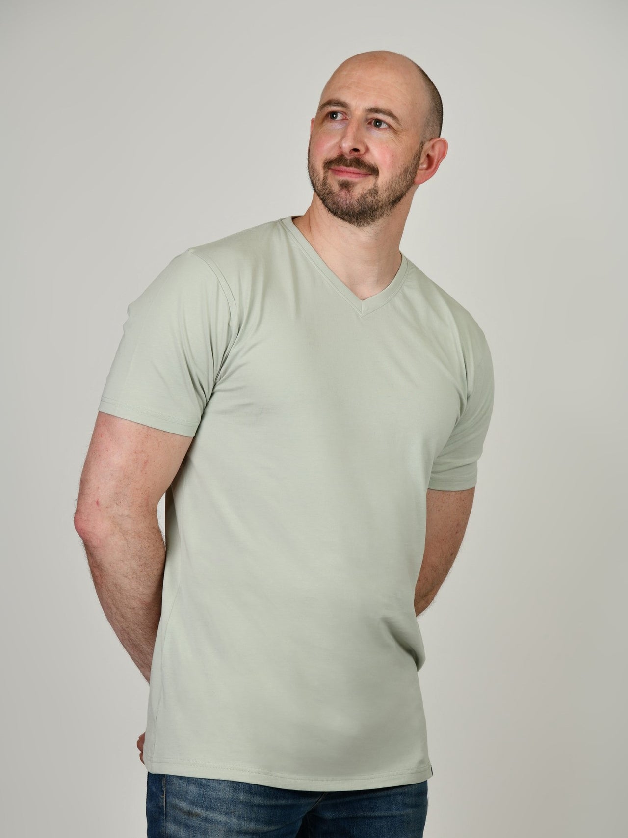 A tall and slim guy in the studio, hands behind back and wearing a sage green XL tall slim v-neck t-shirt.
