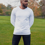 A tall athletic guy wearing a long sleeve white tall t-shirt and smiling in a park with hands behind back.