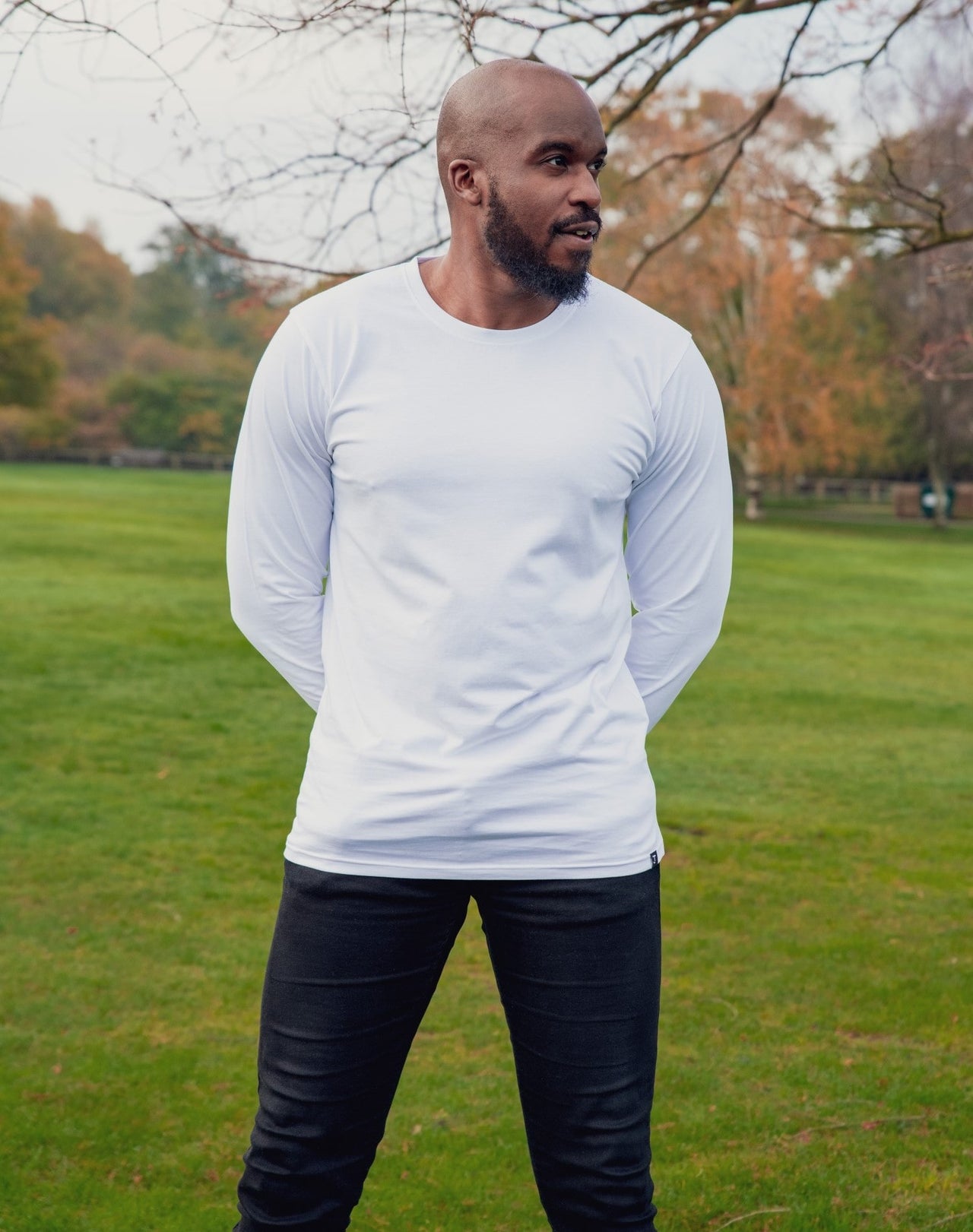 A tall athletic guy wearing a long sleeve white tall t-shirt and smiling in a park with hands behind back.
