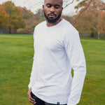 A tall athletic guy wearing a long sleeve white tall t-shirt in a park with one hand in pocket and looking down at the t-shirt logo.