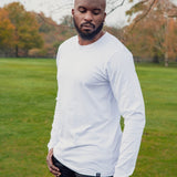 A tall athletic guy wearing a long sleeve white tall t-shirt in a park with one hand in pocket and looking down at the t-shirt logo.