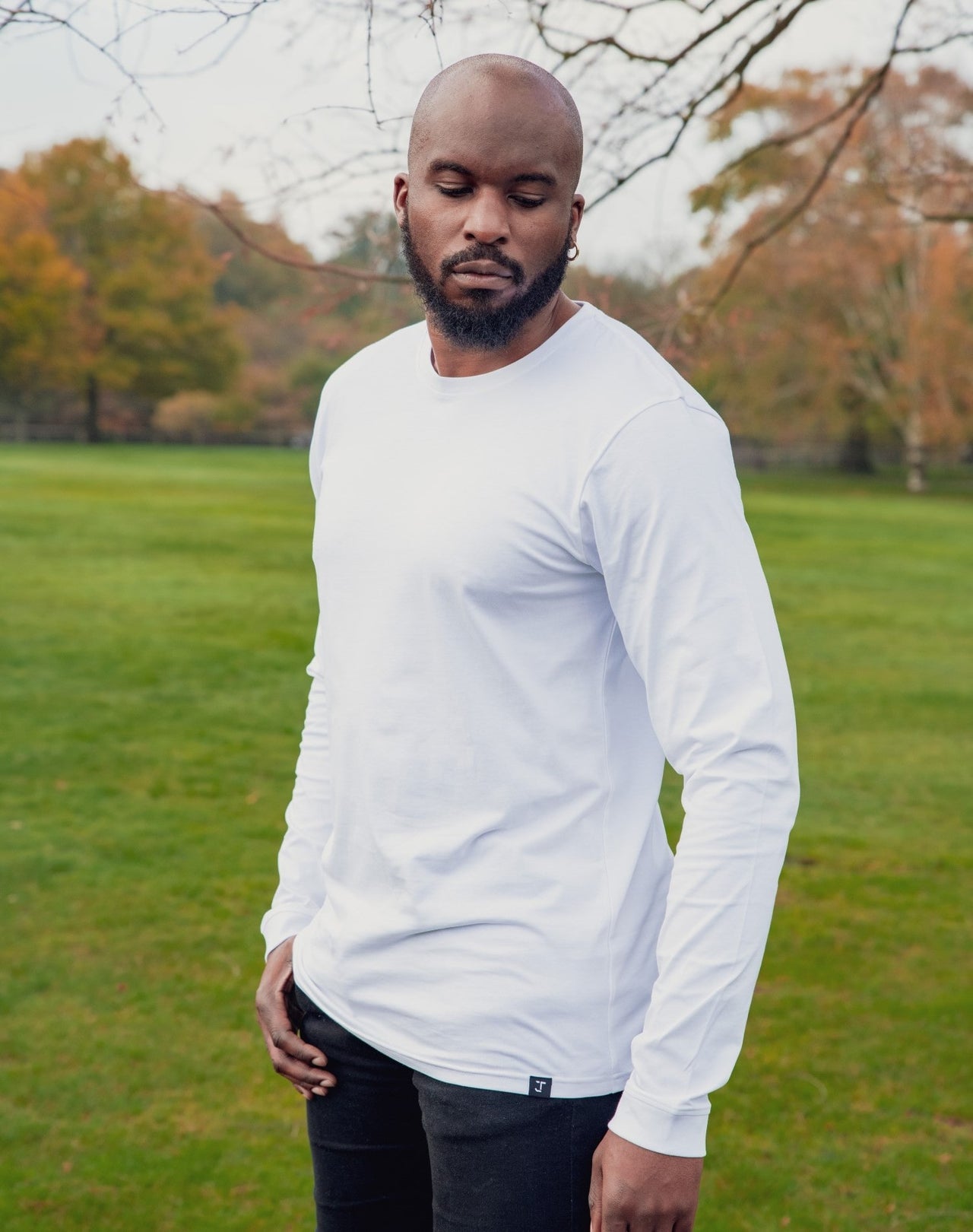 A tall athletic guy wearing a long sleeve white tall t-shirt in a park with one hand in pocket and looking down at the t-shirt logo.
