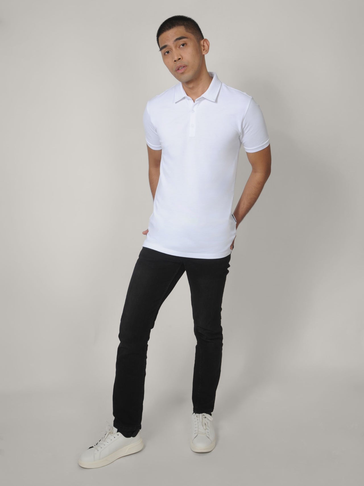 A head to toe shot of a tall skinny guy wearing a tall white pique polo shirt, hands behind back.